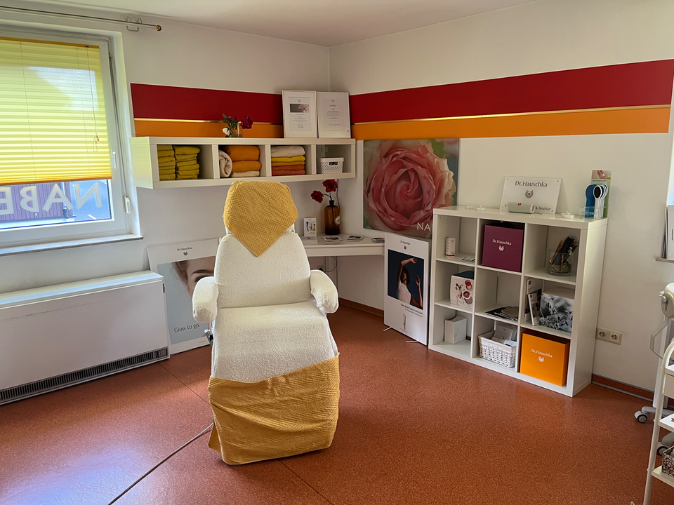 Behandlungsraum in einer Kosmetik- oder Wellnesspraxis. Im Zentrum steht ein weißer Behandlungsstuhl mit gelber Decke und gelbem Tuch über der Kopfstütze. An der Wand befinden sich weiße Regale mit Handtüchern in Gelb- und Orangetönen, Produktboxen und Dekoration. Zwei große Poster, darunter eines mit einer Rose und eines mit einem Frauenporträt, schmücken die Wand. Die Fenster haben gelbe Jalousien, und auf dem Boden liegt ein rötlich-brauner Belag.